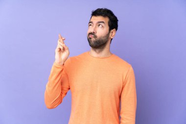 Kafkasyalı yakışıklı adam, izole edilmiş mor arka planda parmakları çapraz ve en iyisini diliyor.