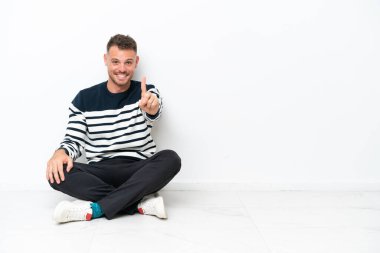 Young man sitting on the floor isolated on white background showing and lifting a finger