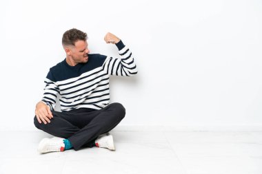 Yerde oturan genç adam beyaz arka planda izole bir şekilde güçlü bir jest yapıyor.