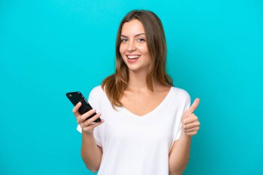 Genç beyaz kadın mavi arka planda izole edildi. Cep telefonuyla onay verdi.