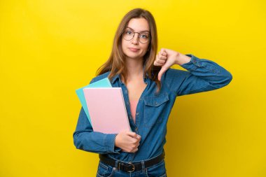 Sarı arka planda izole edilmiş genç beyaz kadın, olumsuz bir ifadeyle baş parmağını aşağı gösteriyor.