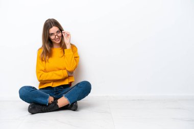 Beyaz arka planda, gözlüklü ve mutlu bir şekilde yerde oturan beyaz tenli bir kadın.