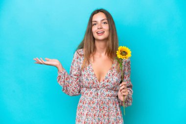 Elinde ayçiçeği tutan beyaz bir kadın mavi arka planda izole edilmiş ve şok olmuş bir yüz ifadesi var.