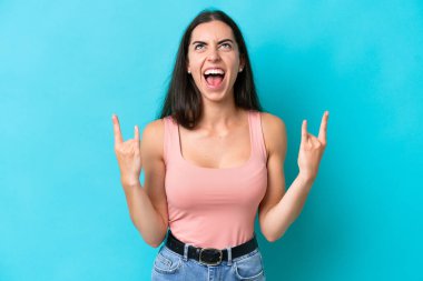 Mavi arka planda izole edilmiş genç beyaz kadın boynuz hareketi yapıyor.