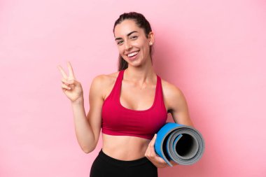 Genç beyaz kadın yoga derslerine gidiyor pembe arka planda izole edilmiş bir minderi tutarken gülümsüyor ve zafer işareti gösteriyor.