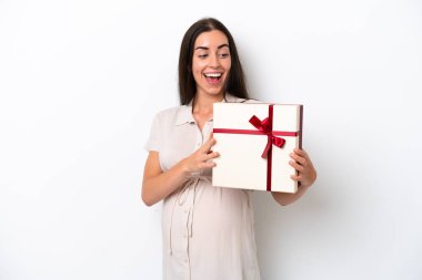 Beyaz arka planda izole edilmiş genç beyaz bir kadın hamile ve elinde şaşırtıcı bir ifadeyle bir hediye tutuyor.