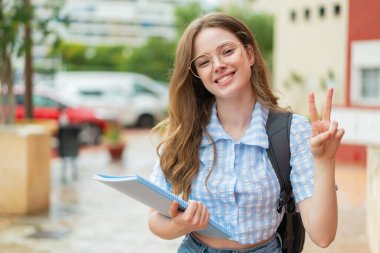 Genç kızıl saçlı bayan açık havada gülümsüyor ve zafer işareti gösteriyor.