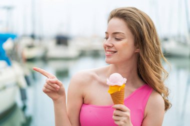 Açık havada kornet dondurmalı genç kızıl saçlı kadın bir ürün sunmak için kenarı işaret ediyor.