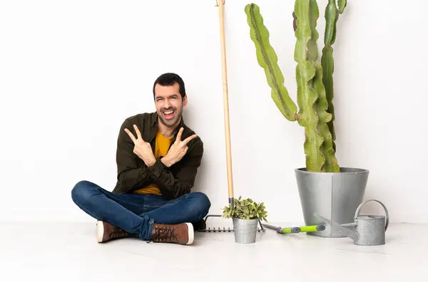 Gardener man sitting on the floor at indoors smiling and showing victory sign