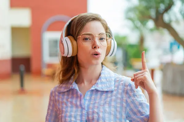 Young Redhead Girl Headphones Outdoors Thinking Idea Pointing Finger — Stock Photo, Image