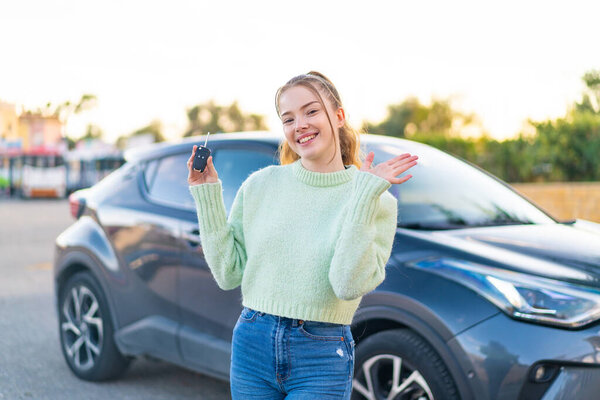 Young pretty girl holding car keys at outdoors saluting with hand with happy expression