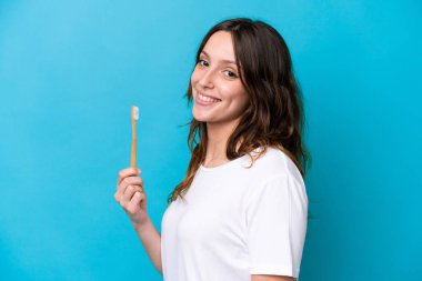 Mavi arka planda izole edilmiş dişlerini fırçalayan beyaz kadın çok gülümsüyor.