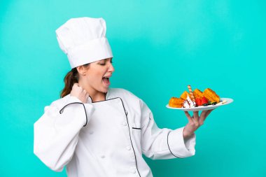 Mavi arka planda zafer kutlamaları yapan Kafkasyalı genç aşçı kadın waffle tutuyor.