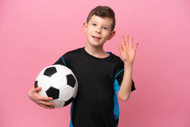 Küçük beyaz futbolcu çocuk pembe arka planda izole edilmiş mutlu bir ifadeyle selamlıyor.