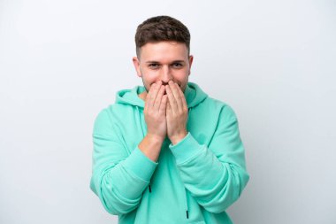 Beyaz arka planda izole edilmiş genç beyaz bir adam mutlu ve gülümseyen elleriyle ağzını kapatıyor.