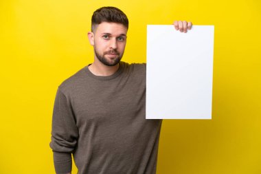 Kafkasyalı genç adam sarı arka planda izole edilmiş boş bir levha tutuyor ve ona bakıyor.