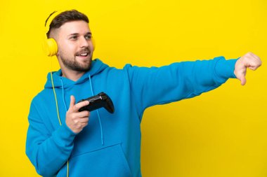 Sarı arka planda izole edilmiş bir bilgisayar oyunu kontrolörüyle oynayan genç beyaz bir adam işaret veriyor.