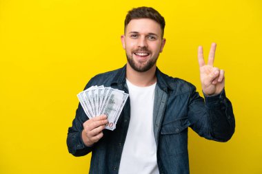 Kafkasyalı genç adam sarı arka planda izole edilmiş bir sürü para alıyor gülümsüyor ve zafer işareti gösteriyor.
