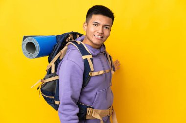 Sarı arka planda izole edilmiş büyük sırt çantalı genç dağcı, davet edildiği için ellerini yana uzatıyor.