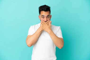 Mavi arka planda izole edilmiş genç beyaz adam elleriyle ağzını kapatıyor.