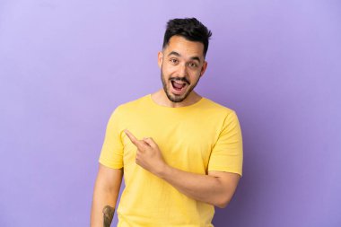 Mor arka planda izole edilmiş genç beyaz adam şaşırmış ve işaret ediyor.