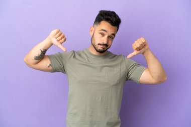 Mor arka planda izole genç beyaz adam baş parmağını iki eliyle aşağı gösteriyor.