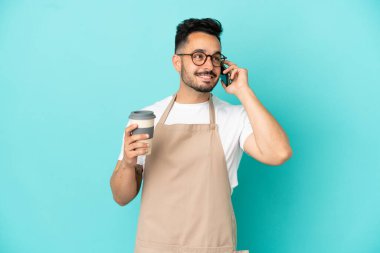 Restoran garsonu Kafkasyalı adam mavi arka planda izole edilmiş. Elinde kahve ve cep telefonu var.