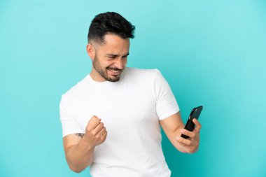 Genç beyaz adam mavi arka planda izole edilmiş cep telefonu kullanıyor ve zafer jesti yapıyor.