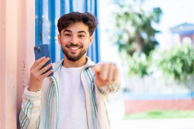 Açık havada cep telefonu kullanan yakışıklı bir Arap, kendine güvenen bir ifadeyle seni işaret ediyor.