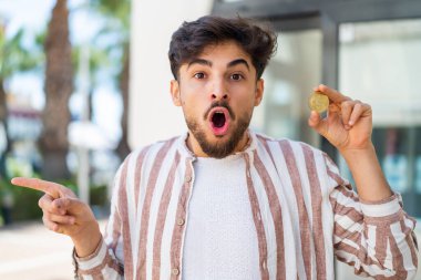 Açık havada Bitcoin tutan yakışıklı bir Arap şaşırmış ve işaret ediyor.