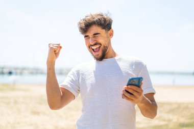 Açık havada telefonu zafer pozisyonunda olan yakışıklı bir Arap.