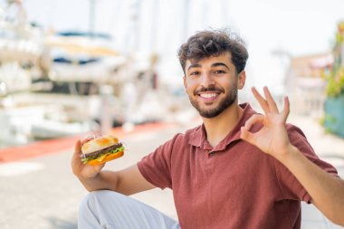 Genç Arap yakışıklı bir adam açık havada hamburger tutuyor. Parmaklarıyla tamam işareti gösteriyor.