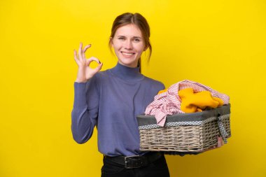Sarı arka planda izole edilmiş çamaşır sepetini tutan genç İngiliz kadın parmaklarıyla tamam işareti gösteriyor.