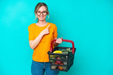 Blonde English young girl holding a shopping basket full of food isolated on blue background pointing to the side to present a product