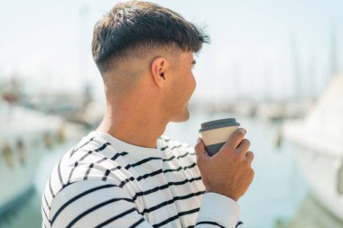 Dışardaki Latin kökenli genç adam elinde kahve tutuyor.