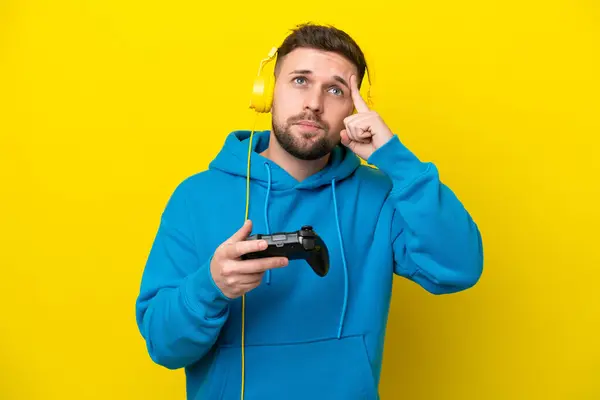 黄色い背景に隔離されたビデオゲームコントローラーで遊ぶ若い白人男性は疑問と思考を持っています — ストック写真