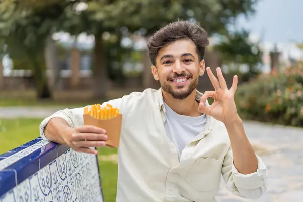 若いアラビアのハンサムな男は 指でOkサインを示す屋外で揚げチップを保持しています — ストック写真