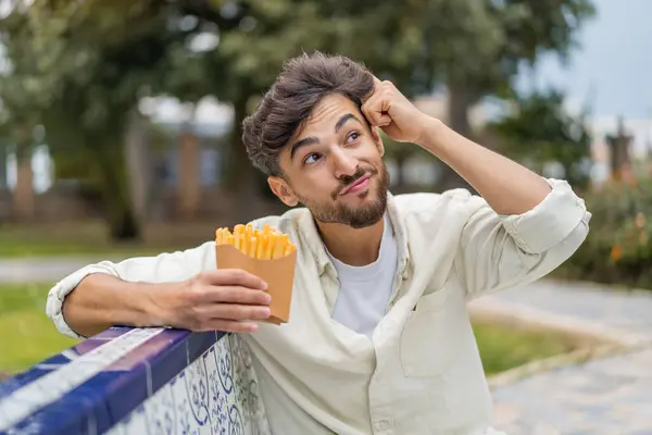 若いアラビアのハンサムな男は疑問を持って 混乱した顔の表情で屋外で揚げチップを持っています — ストック写真