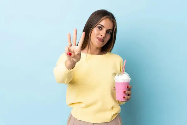 年轻的乌拉圭女人 草莓奶昔与蓝色背景隔离 快乐地数着三根手指 — 图库照片