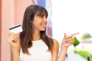 Açık havada kredi kartı tutan genç bir kadın bir ürünü sunmak için kenarı işaret ediyor.