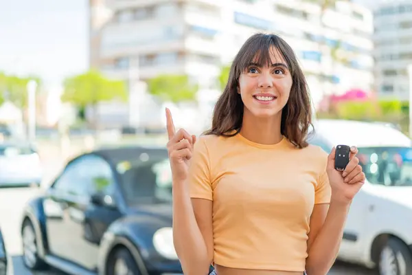 指を上げながら解決策を実現しようとするアウトドアで車の鍵を握っている若い女性 — ストック写真