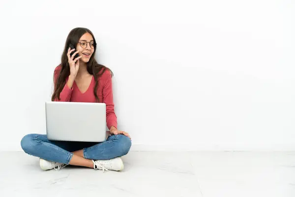 stock image Young French girl sitting on the floor with her laptop keeping a conversation with the mobile phone with someone