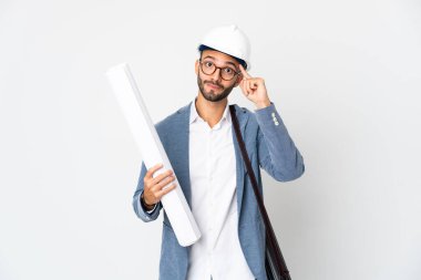 Miğferli ve elinde planları olan genç bir mimar beyaz arka planda bir fikir düşünüyor.