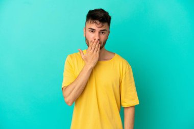 Genç beyaz adam mavi arka planda izole edilmiş, ağzı elle kapatılmış.