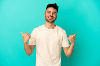 Kafkasyalı genç adam mavi arka planda izole edilmiş. Baş parmakları havada ve gülümsüyor.