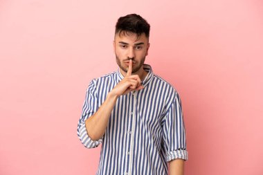 Pembe arka planda izole edilmiş genç beyaz adam sessiz bir jest işareti gösteriyor. Parmağını ağzına sokuyor.