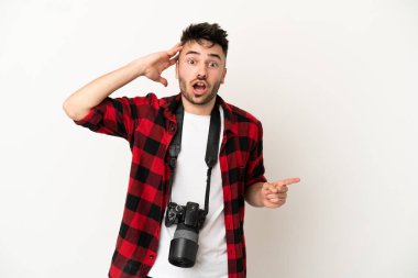Beyaz arka planda izole edilmiş genç fotoğrafçı şaşırmış ve parmağıyla kenarı işaret ediyor.