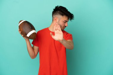 Mavi arka planda ragbi oynayan genç beyaz adam dur hareketi yapıyor ve hayal kırıklığına uğruyor.