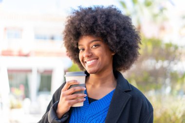 Afro-Amerikalı bir kız açık havada elinde neşeli bir ifadeyle kahve tutuyor.