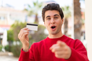 Açık havada elinde kredi kartı tutan yakışıklı genç adam şaşırmış ve ön tarafı işaret ediyor.